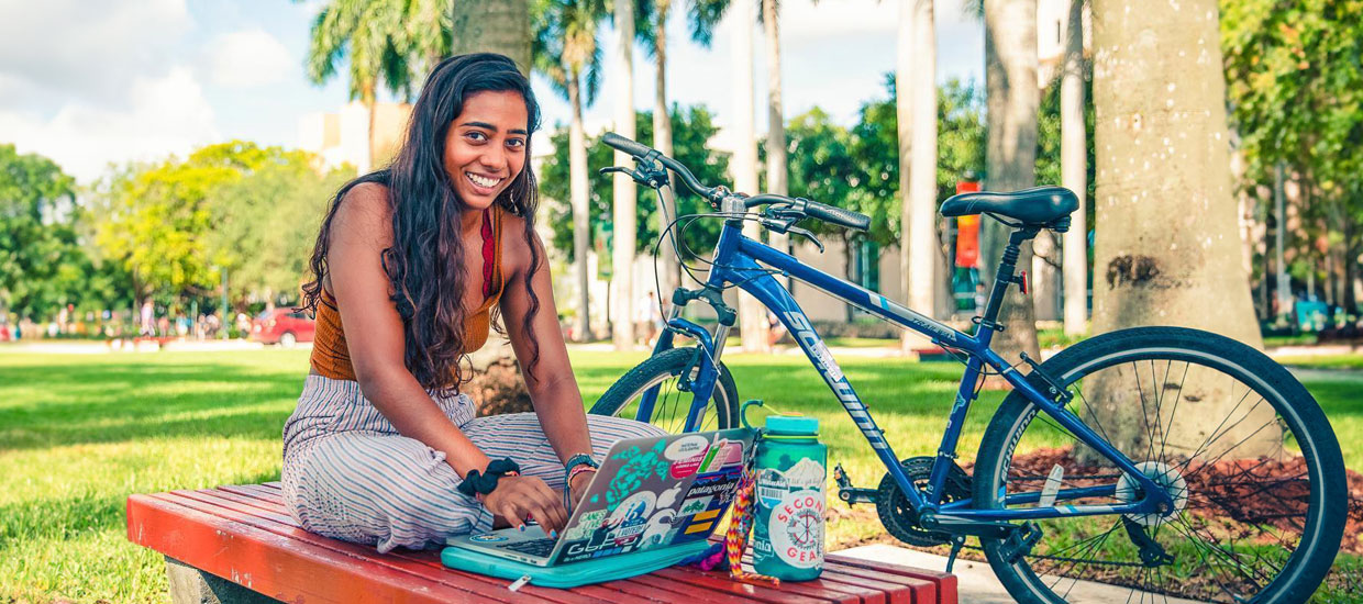 Student sitting outdoors smiling with her laptop and next to a bicycle